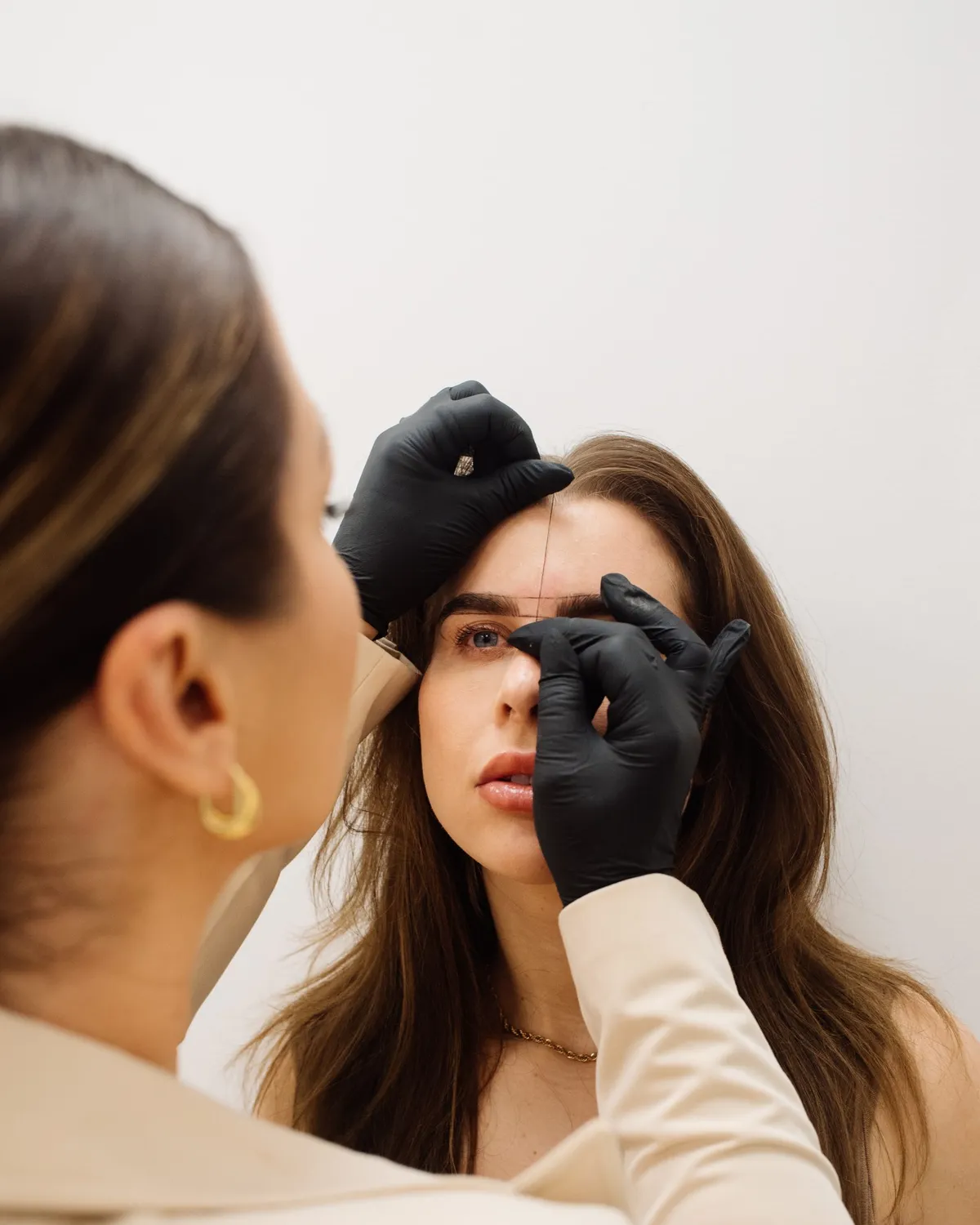 A permanent makeup artist in black gloves mapping a client’s brows with thread